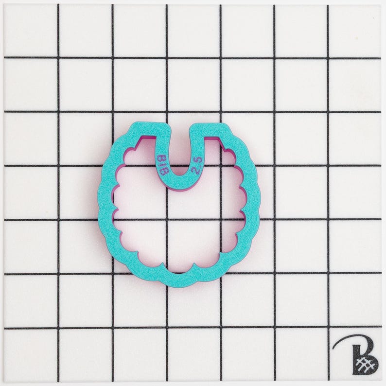 May include: A teal and purple cookie cutter shaped like a bib, with a scalloped edge. The text "BIB" and "2.5" are visible. The cutter is on a white surface with a black grid pattern.