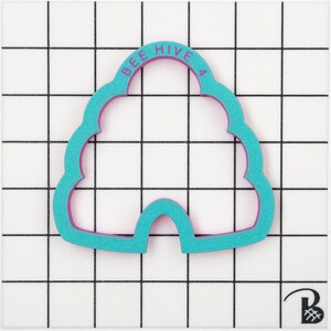 May include: A teal and purple cookie cutter shaped like a beehive. The words "BEE HIVE 4" are printed on the top. The cutter is on a white surface with a black grid pattern.