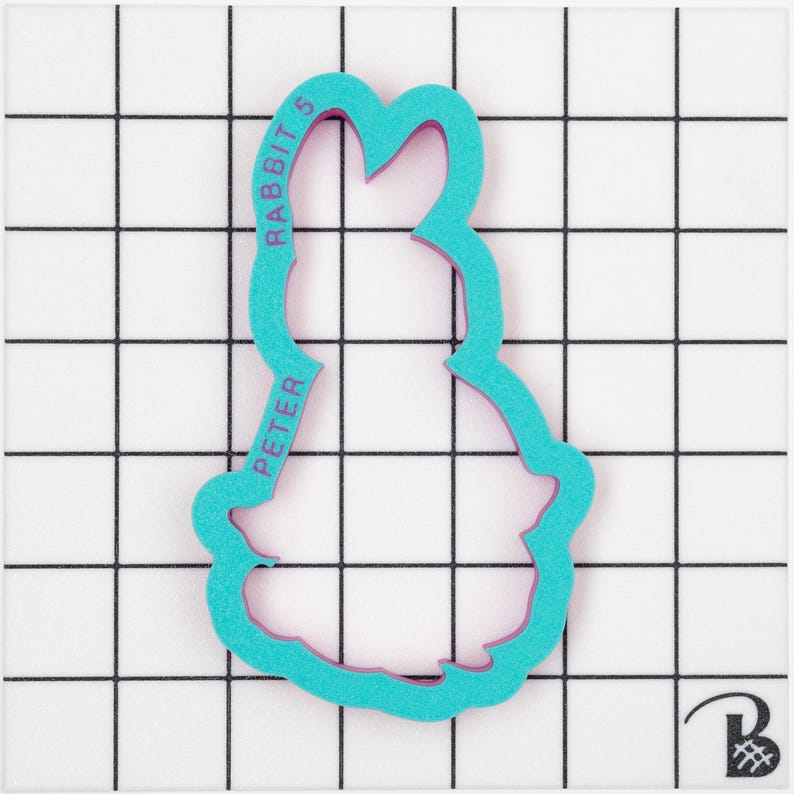 May include: A teal-colored cookie cutter in the shape of a rabbit. The cutter has a pink interior and the text "PETER RABBIT 5" printed vertically along the side. The cutter is on a white surface with a black grid pattern.