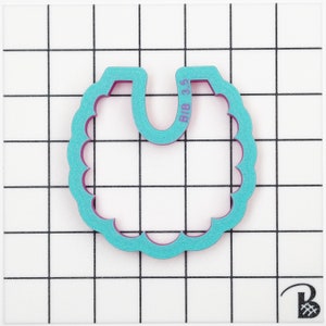 May include: A teal and purple bib-shaped cookie cutter with scalloped edges. The text "BIB 3.5" is printed on the top right side of the cutter. The cutter is on a white surface with a black grid pattern.