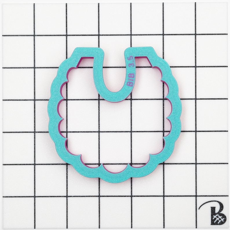 May include: A teal and purple bib-shaped cookie cutter with scalloped edges. The text "BIB 3.5" is printed on the top right side of the cutter. The cutter is on a white surface with a black grid pattern.