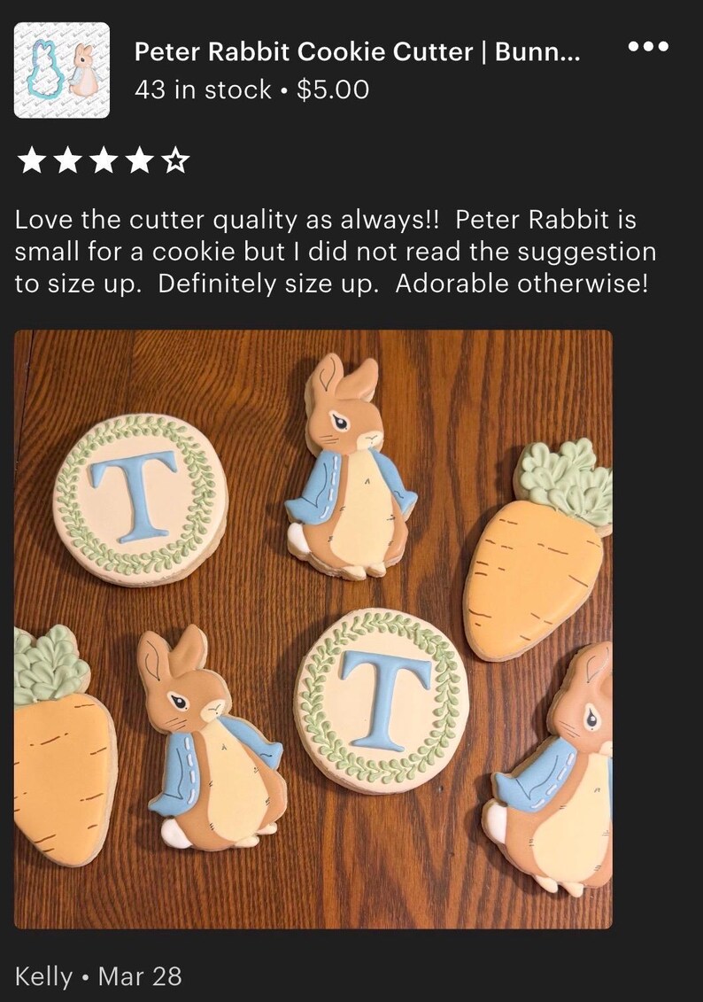 May include: Cookies decorated with Peter Rabbit and carrot designs. The cookies are beige, brown, and blue, with green accents. Some cookies have the letter "T" in blue. The cookies are arranged on a wooden surface.