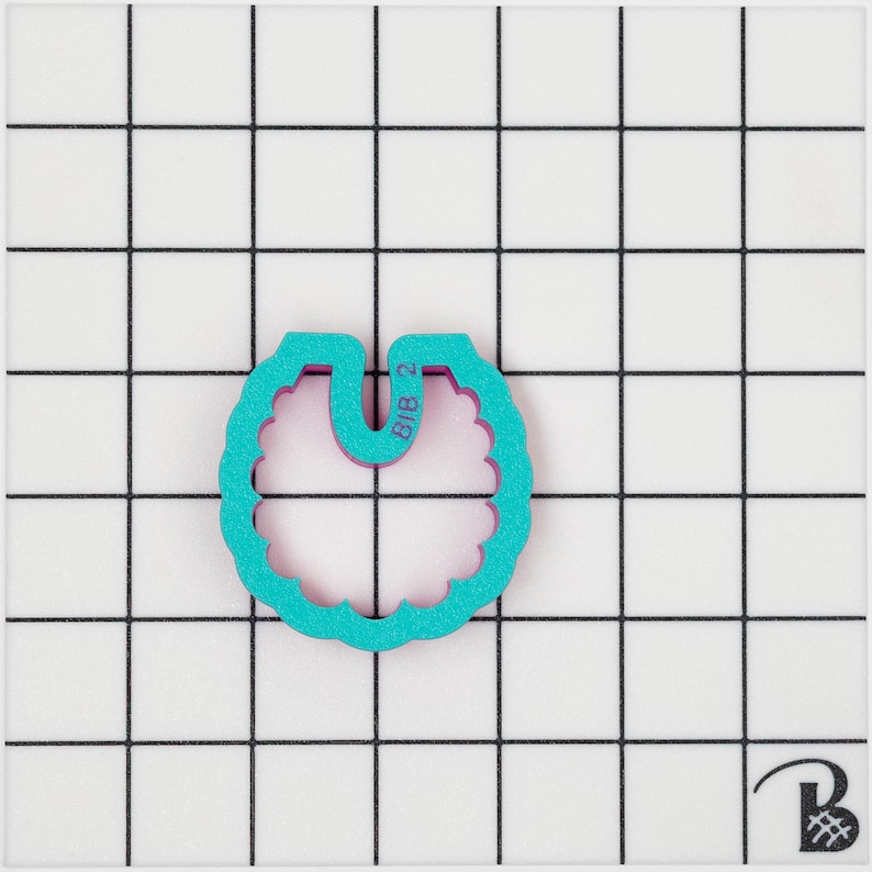 May include: A teal-coloured, scalloped-edge cookie cutter in the shape of a bib, with a purple interior. The text "BIB 2" is visible on the top. The cutter is set against a white grid background.