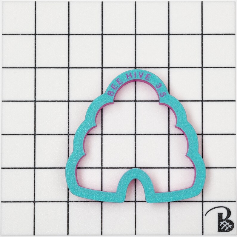 May include: A teal bee hive shaped cookie cutter with a purple interior. The text "BEE HIVE 35" is printed on the top arch of the cutter. The cutter is on a white surface with a black grid pattern.