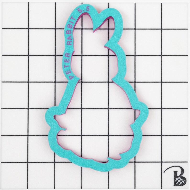 May include: A teal and purple Peter Rabbit cookie cutter. The text "PETER RABBIT 5.5" is printed on the side. The cutter is on a white surface with a black grid pattern.