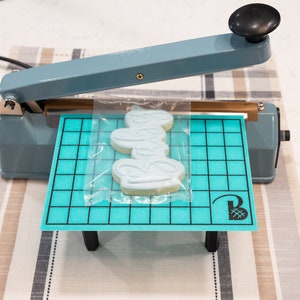 May include: A blue heat sealer with a clear plastic bag containing a light blue cookie with a design on it. The cookie is on a blue and black cutting mat with a logo on it.