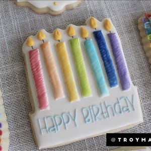 K&ouml;nnte beinhalten: Ein dekorierter Zuckerkeks mit einem Regenbogen aus Kerzen und dem Text "Happy Birthday".