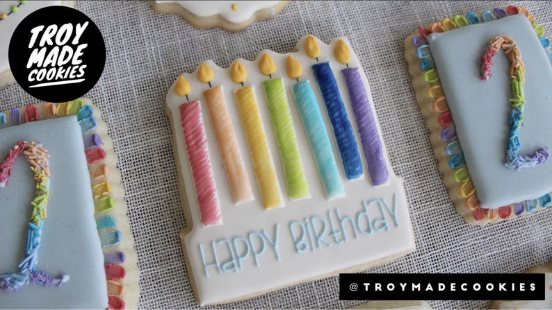 K&ouml;nnte beinhalten: Ein dekorierter Zuckerkeks mit einem Regenbogen aus Kerzen und dem Text "Happy Birthday".