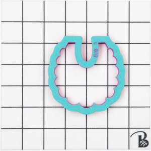 May include: A teal bib-shaped cookie cutter with a scalloped edge and the text "BIB 3" printed on it. The cutter has a purple interior and is set against a white grid background.