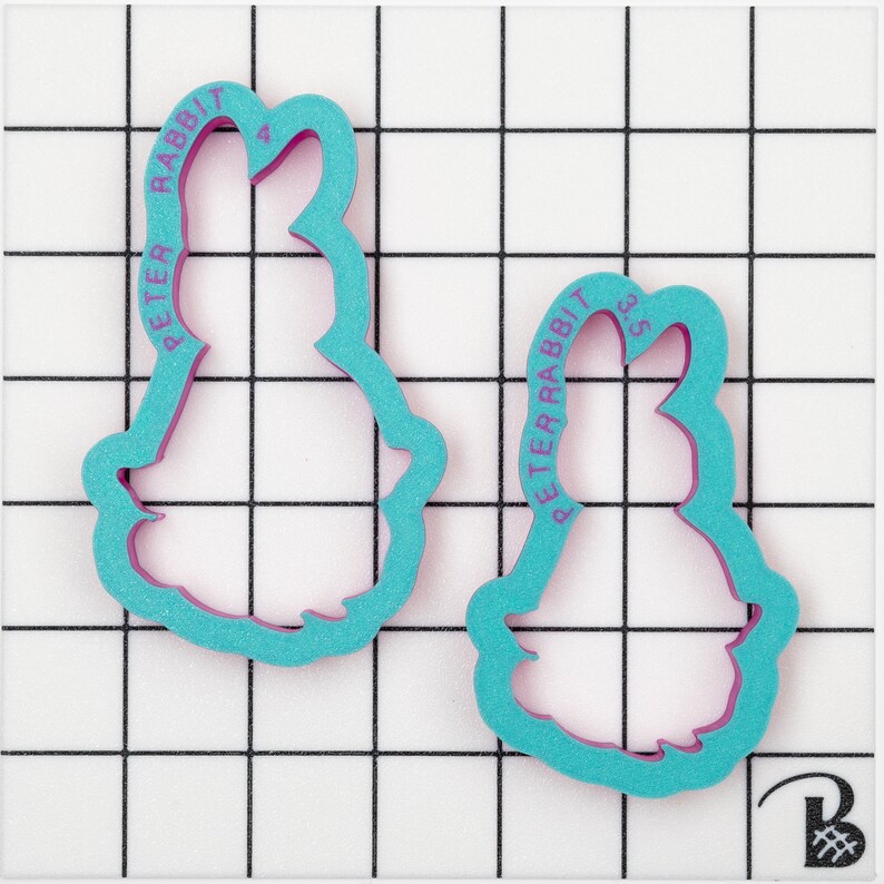 May include: Two Peter Rabbit cookie cutters. The cutters are teal with a pink interior and feature the text "PETER RABBIT" along the side. One cutter is labeled "4" and the other "3.5".