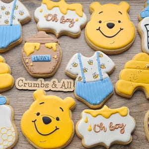 May include: A selection of decorated biscuits in the shape of a baby onesie, a honey pot, a beehive, and a baby bottle. The biscuits are decorated with yellow, blue, white, and brown icing. Some biscuits have the words "Oh boy" and "Hunny".
