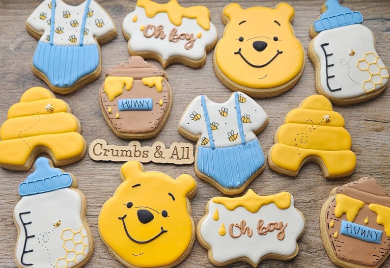 May include: A selection of decorated biscuits in the shape of a baby onesie, a honey pot, a beehive, and a baby bottle. The biscuits are decorated with yellow, blue, white, and brown icing. Some biscuits have the words "Oh boy" and "Hunny".