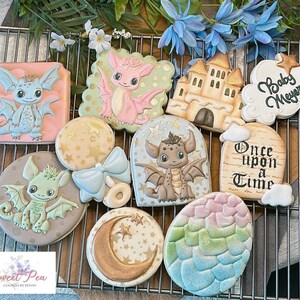 May include: A set of ten decorated sugar cookies featuring a baby dragon theme. The cookies are decorated with pastel colours, gold accents, and various designs including a dragon, a castle, a star, a moon, a bow, and a sign that reads "Once Upon a Time".