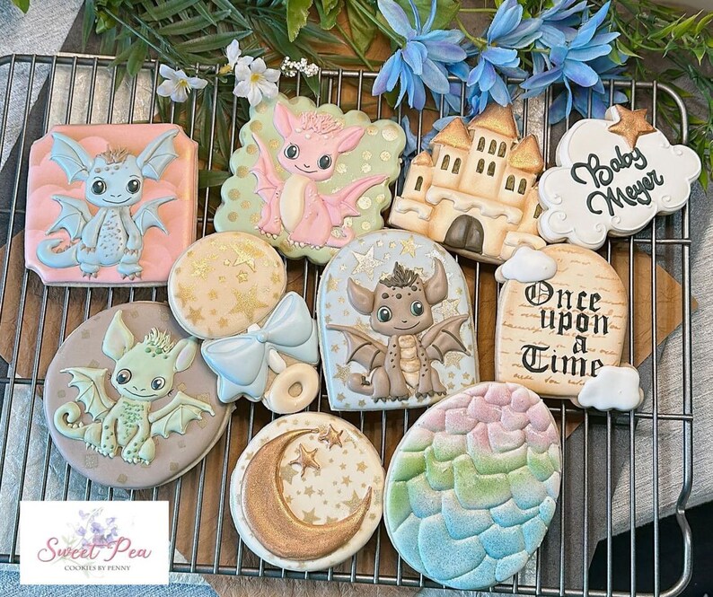 May include: A set of ten decorated sugar cookies featuring a baby dragon theme. The cookies are decorated with pastel colours, gold accents, and various designs including a dragon, a castle, a star, a moon, a bow, and a sign that reads "Once Upon a Time".