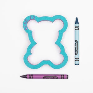 May include: A blue foam bear-shaped cookie cutter with the text "BEAR 3.5" on the side. Two crayons, one blue and one purple, are next to the cookie cutter.