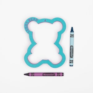 May include: A turquoise blue teddy bear shaped cookie cutter with the words "Teddy Bear 4" around the edge. Two crayons, one blue and one purple, are next to the cookie cutter.