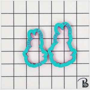 May include: Two teal-colored cookie cutters in the shape of Peter Rabbit, with the text "PETER RABBIT" and "2.5" printed on them. The cutters are on a white grid background.