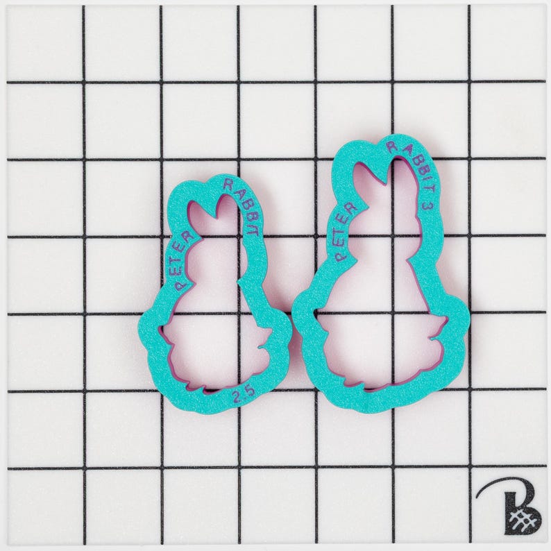 May include: Two teal-colored cookie cutters in the shape of Peter Rabbit, with the text "PETER RABBIT" and "2.5" printed on them. The cutters are on a white grid background.
