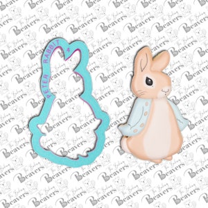 May include: A Peter Rabbit cookie cutter and a decorated cookie. The cookie is light brown with a blue jacket and black eyes. The cutter is light blue and has the words "PETER RABBIT" printed on it.
