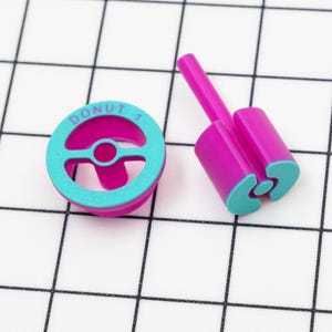 May include: Two pink and blue plastic donut cutters. One cutter is shaped like a donut with a hole in the center. The other cutter is shaped like a donut with a hole in the center and a handle.