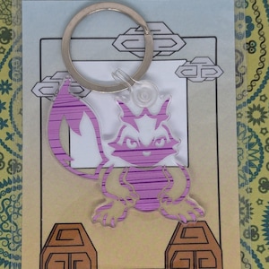 May include: A clear acrylic keychain featuring a purple and white cartoon creature design. The keychain has a silver metal ring and a decorative background with brown and tan elements. The creature has a striped pattern.