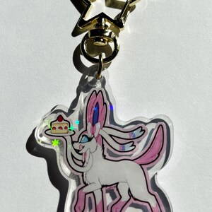 Könnte beinhalten: Ein durchsichtiger Acryl-Schlüsselanhänger mit einer rosa und weißen Comicfigur, die ein Stück Kuchen hält. Der Schlüsselanhänger hat einen goldenen sternförmigen Verschluss.