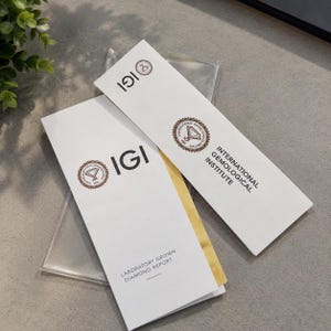 Może przedstawiać: Dwa białe, prostokątne raporty dotyczące diamentów wyhodowanych w laboratorium z International Gemological Institute. Każdy dokument zawiera brązowe, okrągłe logo IGI, skrót IGI oraz pełną nazwę instytutu wydrukowaną czarną czcionką.