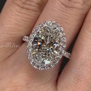 May include: An oval-cut diamond ring with a halo of smaller diamonds. The ring is set in a silver band and sparkles brightly. The image is a close-up, showcasing the intricate details of the jewelry.