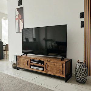 Puede incluir: Un mueble de televisión rectangular con acabado de madera natural, dos armarios laterales y un estante central abierto. La unidad está sostenida por un marco de metal negro e incluye tiradores de metal negro en las puertas de los armarios.