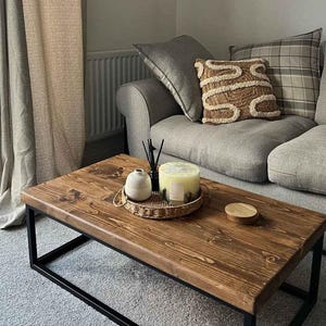 May include: A rectangular coffee table with a dark brown stained wooden top and a black metal frame. On the table sits a small round woven tray containing a white ceramic vase, a black reed diffuser, and a large cream-colored candle.