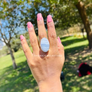 K&ouml;nnte beinhalten: Ein silberner Ring mit einem gro&szlig;en, ovalen, blau-wei&szlig; gestreiften Stein. Der Ring wird auf einer Hand mit rosa Nagellack gezeigt. Der Text "SHUBHJEWELRYSHOP" ist unten im Bild zu sehen.