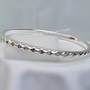 May include: A silver bangle bracelet with a repeating diamond pattern around the band. The bracelet is resting on a white surface. The text "SHUBHJEWELRYSHOP" is visible on the bracelet.