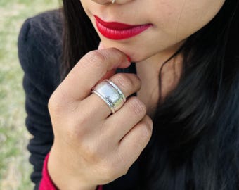 May include: A silver ring with a wide band is worn on a finger. The ring has a smooth, polished surface with a slightly domed top. The person is wearing a black top with a red undershirt and has red lipstick.