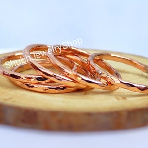 Op de afbeelding: Een verzameling gehamerde koperen ringen, elk met een getextureerd oppervlak, is gerangschikt op een houten schijf. De ringen hebben een warme, roodbruine tint en het licht reflecteert op hun onregelmatige oppervlakken. De ringen zijn op elkaar gestapeld.