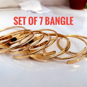 May include: A set of seven gold-colored bangles. Each bangle has a simple, open design with a flat, rectangular accent. The text "SET OF 7 BANGLE" is displayed in red. The jewelry is arranged on a white surface.
