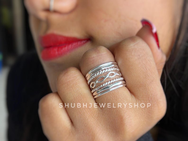 May include: A stackable silver ring set with various designs, including twisted rope and infinity symbols. The rings are worn on a finger, with the text "SHUBHJEWELRYSHOP" visible in the background.