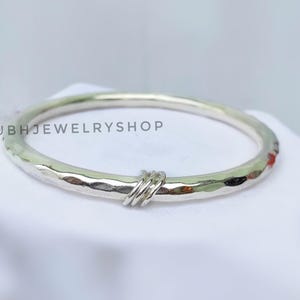 May include: A silver bangle bracelet with a hammered texture and a small, twisted accent. The bracelet is circular and sits on a white surface. The text "SHUBHJEWELRYSHOP" is visible.