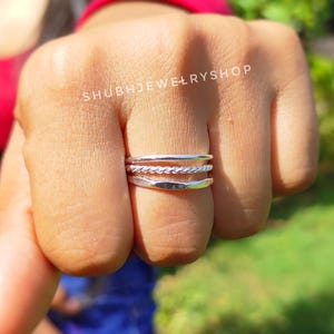 Puede incluir: Tres anillos de plata apilados en un dedo. El anillo superior es liso, el anillo central tiene un diseño de cuerda trenzada y el anillo inferior también es liso. Los anillos se muestran sobre un fondo borroso.