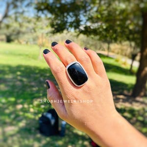 Könnte beinhalten: Ein großer, rechteckiger Ring mit einem schwarzen Edelstein und einem silberfarbenen Rand. Der Ring wird an einer Hand mit schwarzem Nagellack getragen. Der Text "SHUBHJEWELRYSHOP" ist sichtbar.