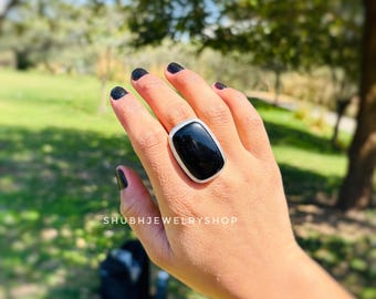 Puede incluir: Un anillo grande, rectangular, con una piedra preciosa negra y un borde plateado. El anillo se lleva en una mano con esmalte de uñas negro. El texto "SHUBHJEWELRYSHOP" es visible.