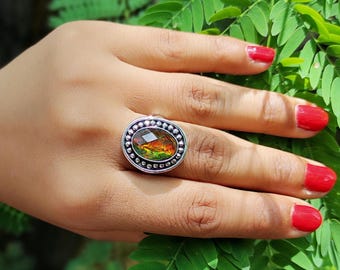 Könnte beinhalten: Ein silberner Ring mit einem ovalen, mehrfarbigen Edelstein in Orange-, Gelb- und Grüntönen. Der Ring ist mit kleinen, runden Verzierungen versehen. Die Hand, mit rotem Nagellack, liegt auf einem grünen Blatt-Hintergrund.