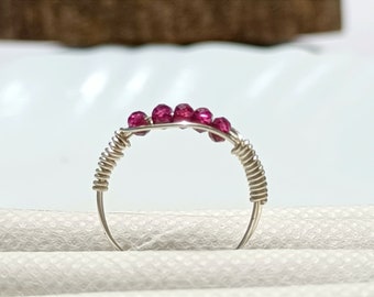 Könnte beinhalten: Ein silberner Drahtring mit einer Reihe kleiner, leuchtend rosa Edelsteine. Die Edelsteine sind über der Oberseite des Rings angeordnet, wobei der Draht zur Dekoration um das Band gewickelt ist. Der Ring wird auf einer weißen Oberfläche gezeigt.