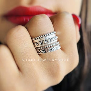 Peut inclure: Une main portant une série de bagues en argent. Les bagues présentent divers motifs, notamment une texture en forme de corde, de petits détails carrés et des motifs floraux. Les bagues sont empilées sur un doigt, mettant en valeur les différents styles.