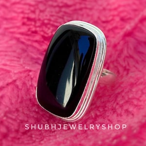 May include: A rectangular black onyx ring with a silver band. The polished black stone is set in a silver bezel with a layered design. The ring is displayed on a pink, textured surface. The text "SHUBHJEWELRYSHOP" is at the bottom.