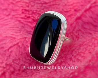Könnte beinhalten: Ein rechteckiger Ring aus schwarzem Onyx mit einem silbernen Band. Der polierte schwarze Stein ist in einer silbernen Lünette mit einem geschichteten Design gefasst. Der Ring wird auf einer rosa, strukturierten Oberfläche präsentiert. Der Text "SHUBHJEWELRYSHOP" befindet sich unten.