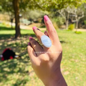 K&ouml;nnte beinhalten: Ein Silberring mit einem gro&szlig;en, ovalen, hellblauen Edelstein. Der Ring befindet sich an einer Hand mit rosa Nagellack, vor einem verschwommenen Hintergrund im Freien. Der Text "SHUBHJEWELRYSHOP" ist sichtbar.