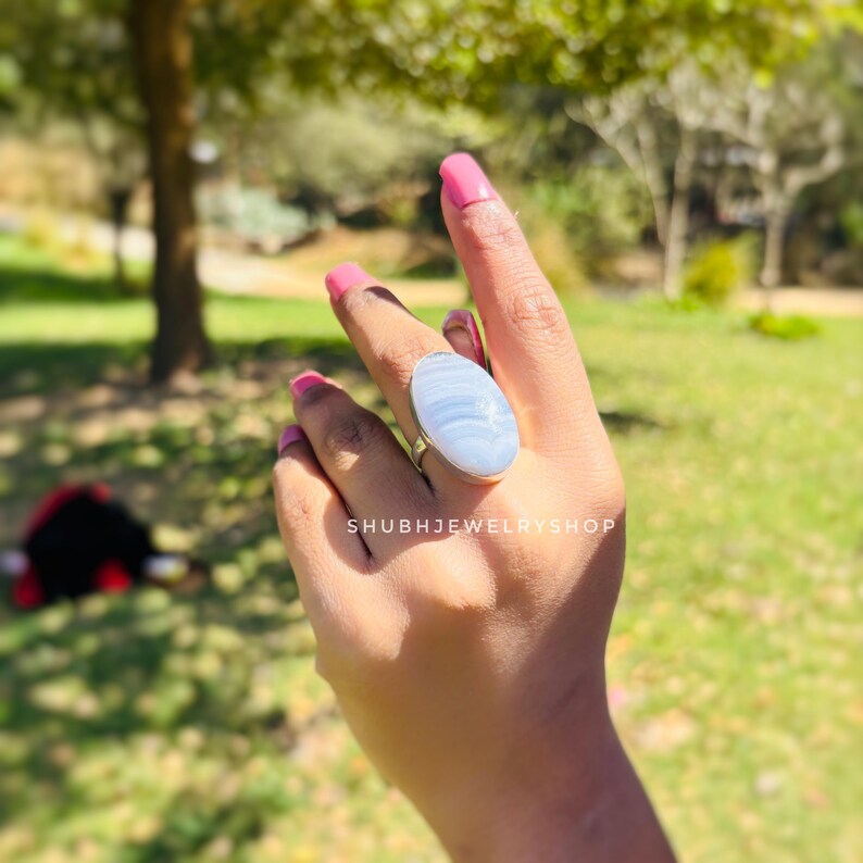K&ouml;nnte beinhalten: Ein Silberring mit einem gro&szlig;en, ovalen, hellblauen Edelstein. Der Ring befindet sich an einer Hand mit rosa Nagellack, vor einem verschwommenen Hintergrund im Freien. Der Text "SHUBHJEWELRYSHOP" ist sichtbar.