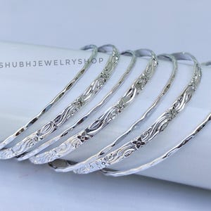 May include: Six silver bangles with floral and textured designs are displayed. The bangles are stacked on a white surface. The text "SHUBHJEWELRYSHOP" is visible on the surface.