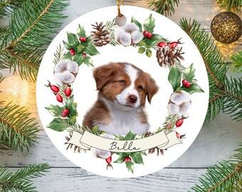 Puede incluir: Un adorno navideño de cerámica blanca con una corona de bolas de algodón, acebo y piñas. Un perro marrón y blanco con el nombre "Bella" está en el centro de la corona.