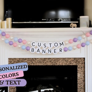 May include: A white banner with the text "CUSTOM BANNER" in black letters. The banner is decorated with a garland of pastel pink, purple, and blue balloons.